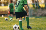 Soccer player in green jersey and socks standing on a field, holding their knee, with other players and soccer balls in the background.